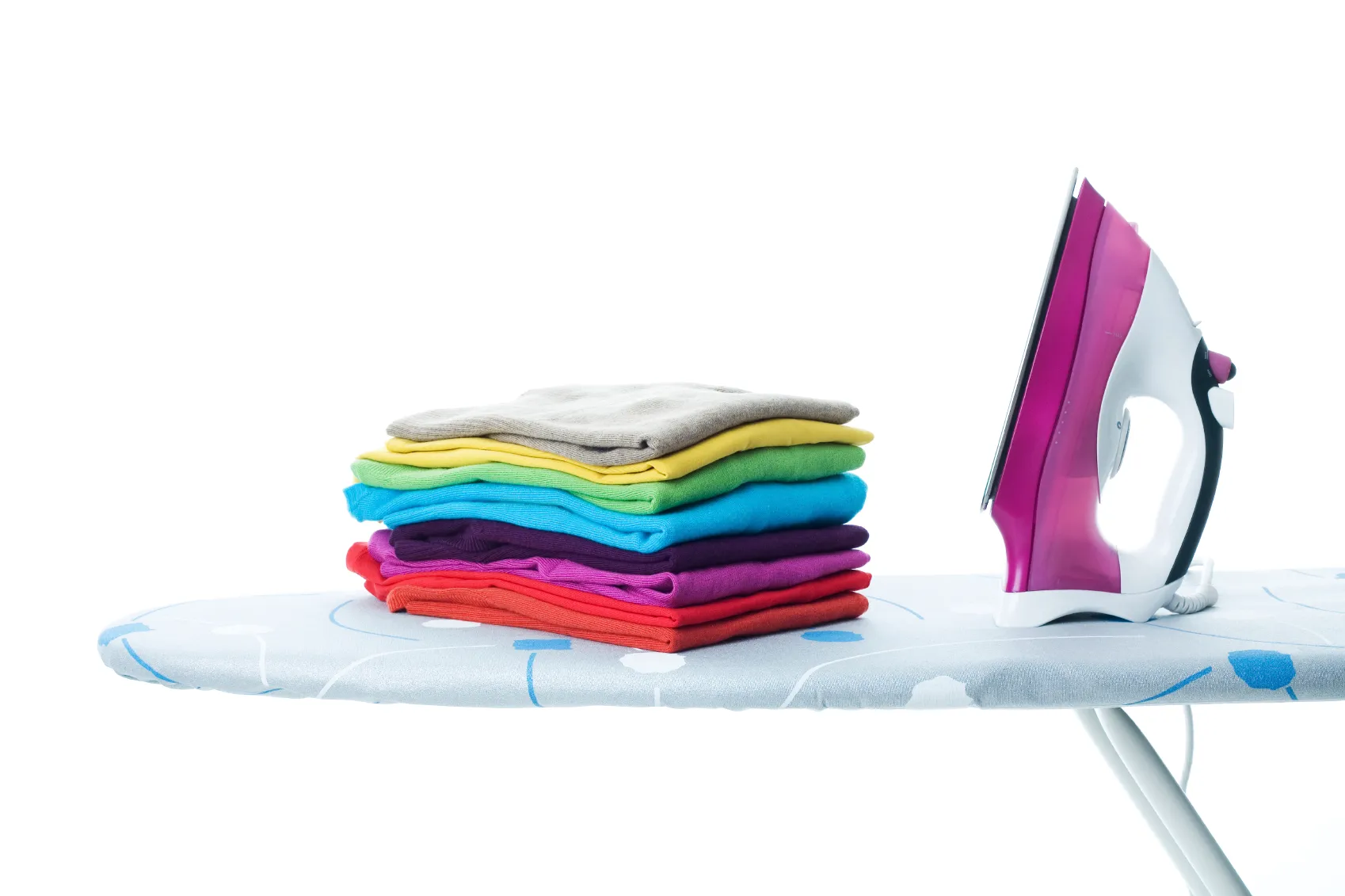 A pink steam iron on an ironing board beside a stack of folded, brightly coloured jumpers